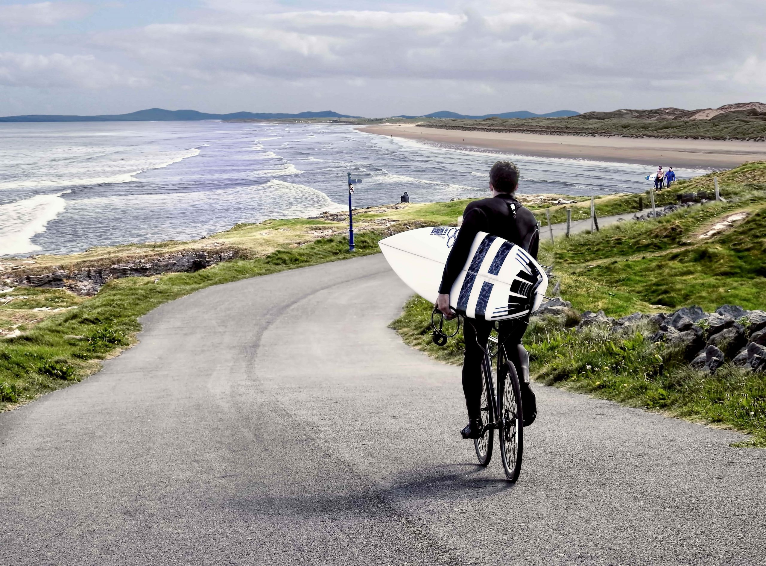 Man cycling down Tullan Strand with surf board under his arm.