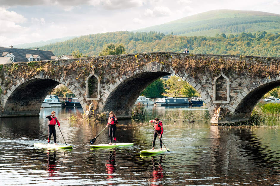 Group of young adults on stand up paddleboards approaching a bridge on River Barrow